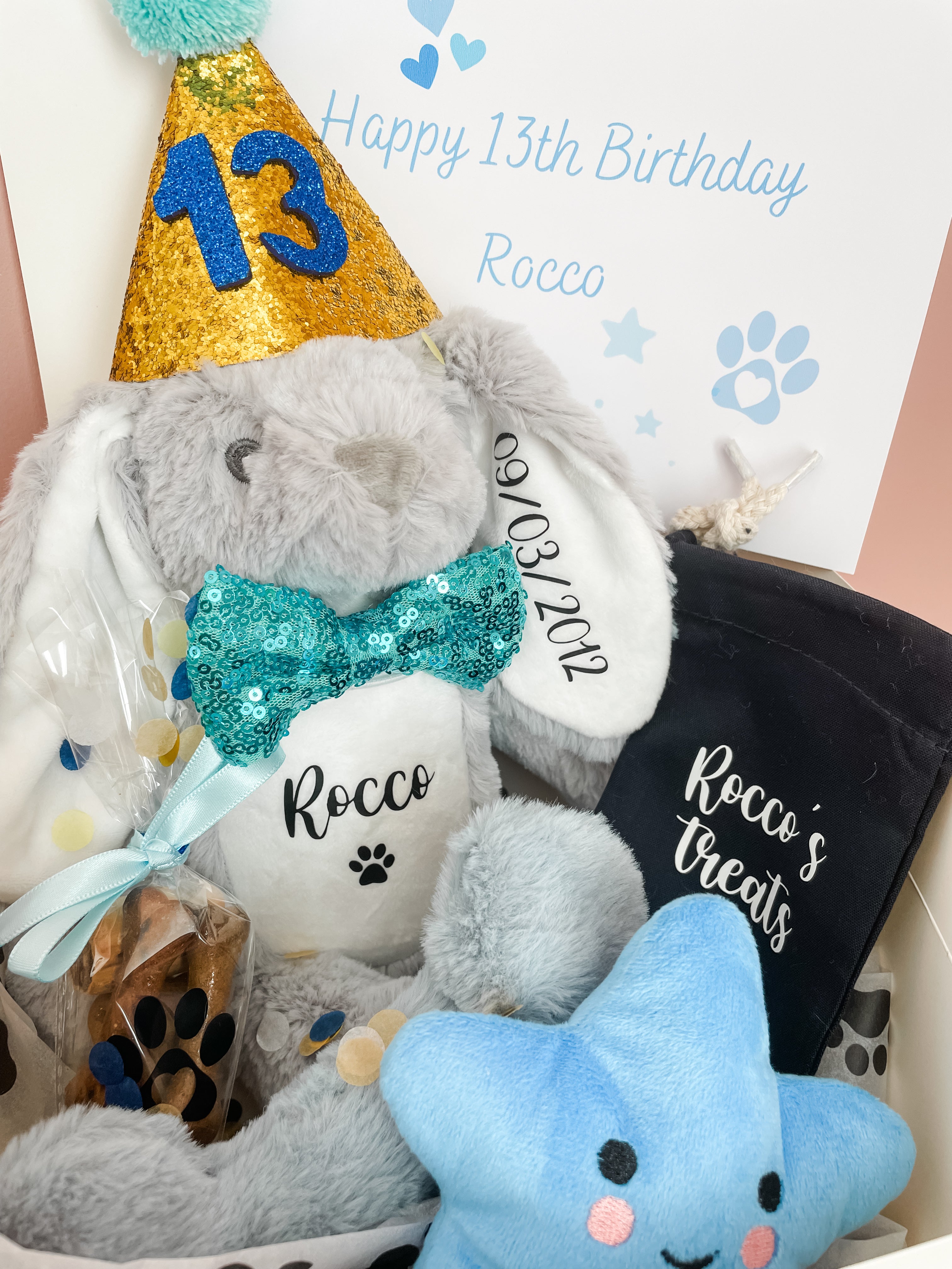 A blue birthday hamper for dogs containing a personalised toy bunny, a fluffy bone toy, a blue star toy, a blue and gold birthday hat, a bow tie, a personalised treat bag and a treat cone with dog treats inside, all packaged in a white gift box with a blue ribbon and a gift message card.