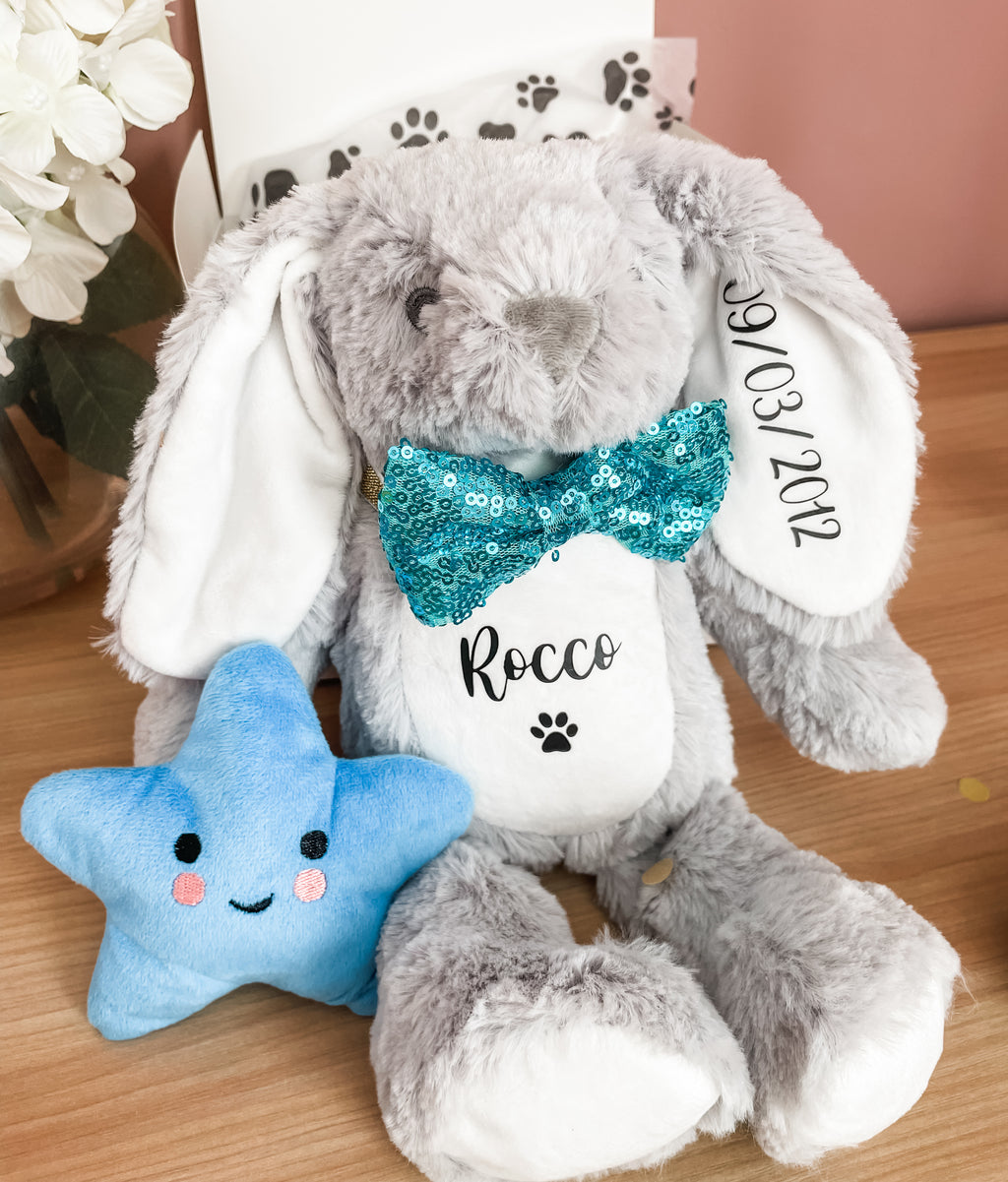 Personalised grey bunny plush toy for dog's with custom name and birthday date placed next to a blue star squeaky toy. Custom dog plush toys.