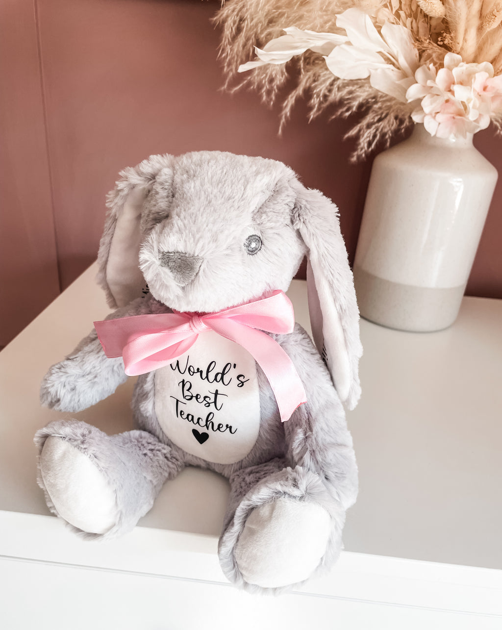 A grey plush bunny toy with long floppy ears, a pink satin bow around its neck, and a white belly patch that reads 'World's Best Teacher' in black script. Personalised bunny keepsake gift for teachers with custom message from student.