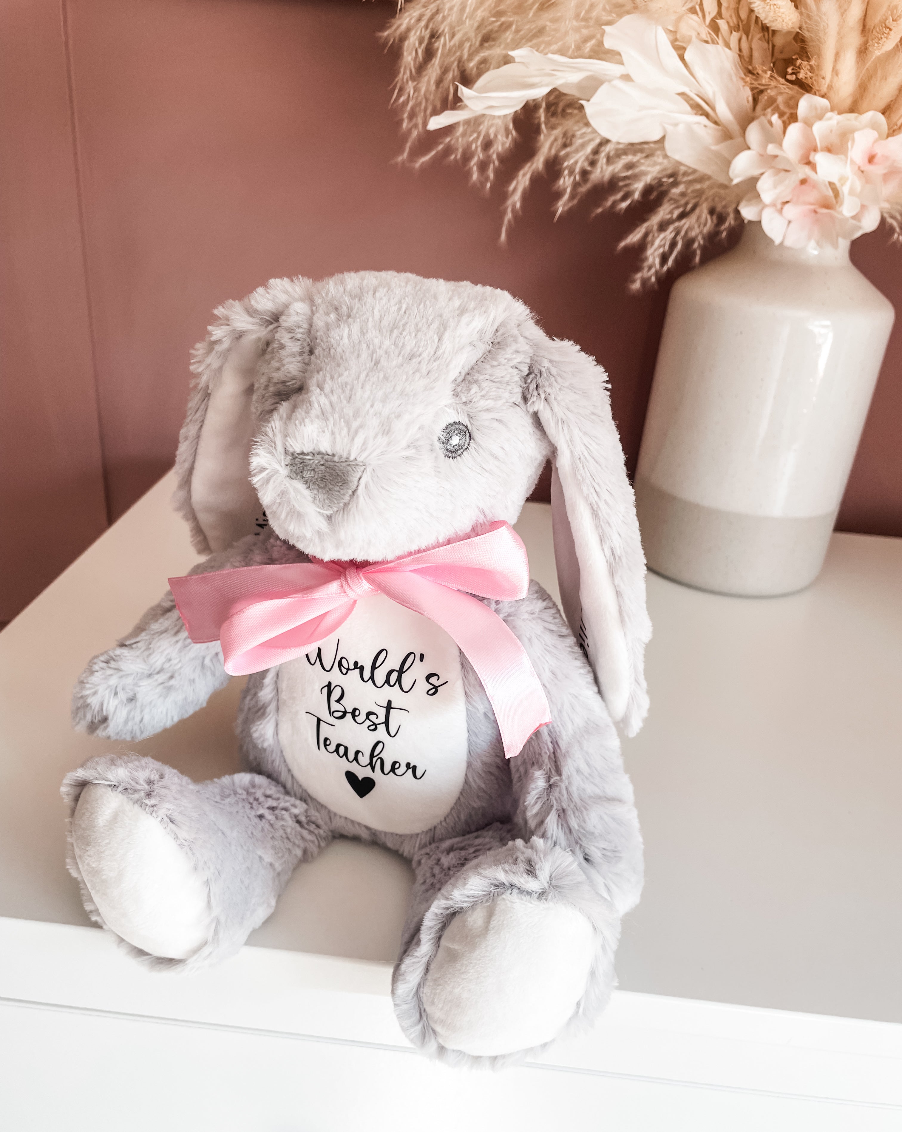 A grey plush bunny toy with long floppy ears, a pink satin bow around its neck, and a white belly patch that reads 'World's Best Teacher' in black script. Personalised bunny keepsake gift for teachers with custom message from student.