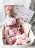 A curated birthday dog gift box filled with pink plush toys and accessories, including a personalised bunny wearing a glittery party hat and bow tie, a star-shaped plush, a bone toy, treat bag and a printed birthday card reading 'Happy Birthday'.