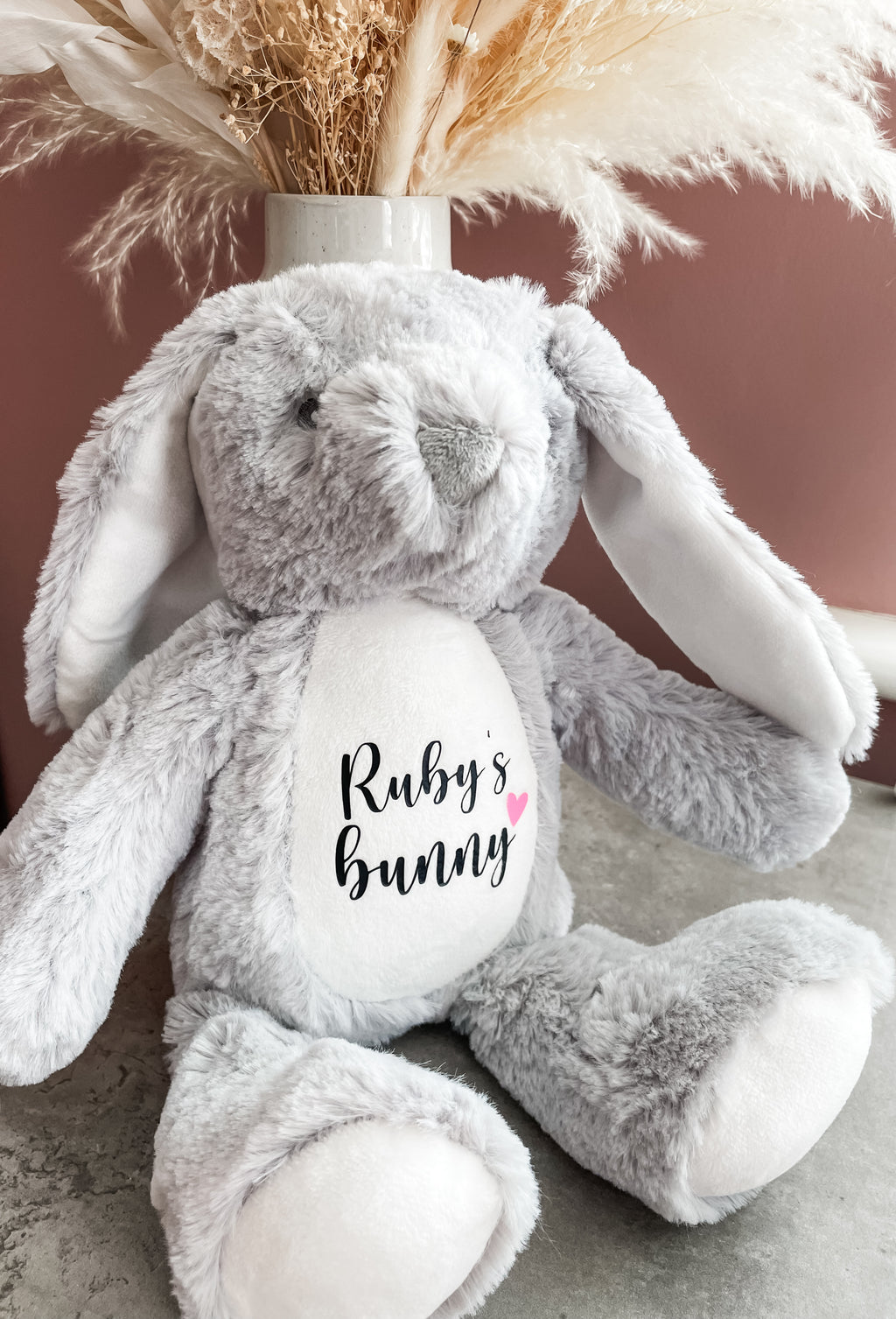 A plush grey bunny stuffed animal with long floppy ears and a white belly patch printed with dog's name and the word 'bunny' with a small heart. The bunny is posed in front of a dusty rose wall with dried pampas grass in a vase behind it.