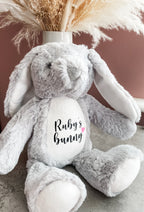 A plush grey bunny stuffed animal with long floppy ears and a white belly patch printed with dog's name and the word 'bunny' with a small heart. The bunny is posed in front of a dusty rose wall with dried pampas grass in a vase behind it.