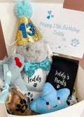 A blue birthday hamper for dogs including a personalised bunny toy, a fluffy bone toy, a blue star toy, a blue and gold birthday hat with a bow tie, a personalised treat bag and a treat cone with various dog treats inside, all packaged in a white gift box with a blue ribbon and a gift message card.