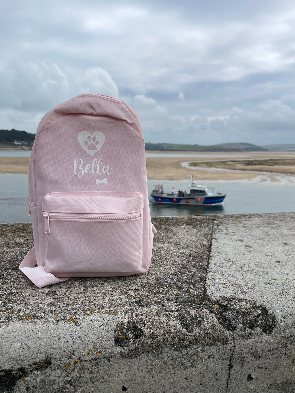 Pink personalised dog backpack at the beach with boat in the background, perfect for dog holidays and adventures.