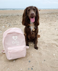 Cocker Spaniel on a sandy beach next to a pink personalised dog backpack with a white paw print and bone design. Custom dog holiday bag.