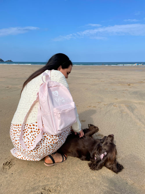 Personalised pink dog walking backpack with custom dog name, carried at the beach beside a Cocker Spaniel.
