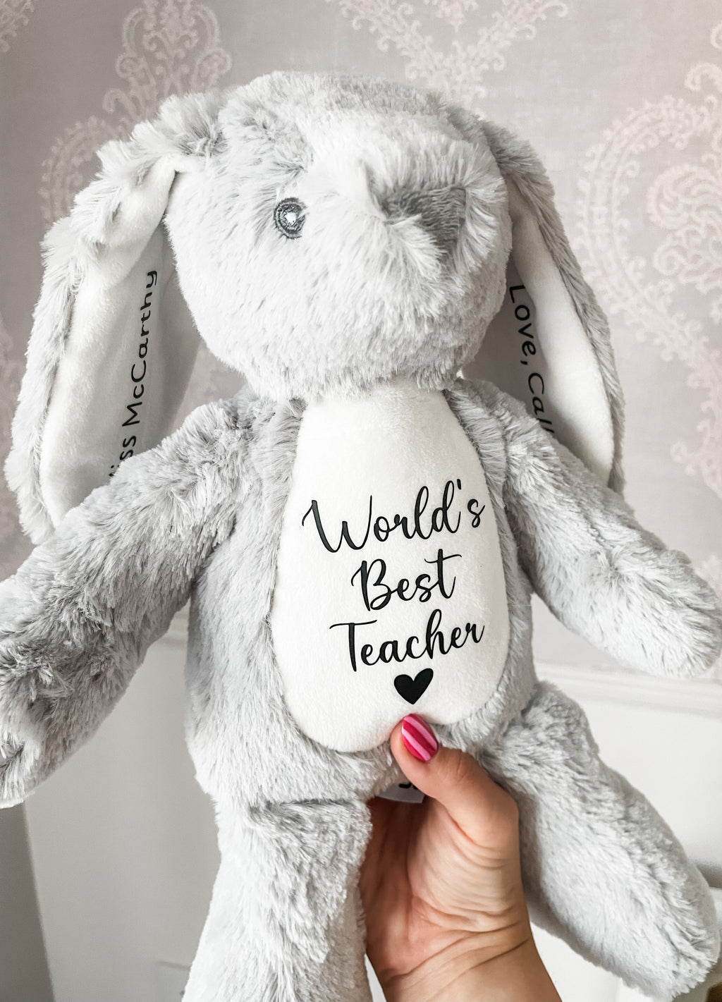 A light grey plush bunny stuffed animal held by a hand, featuring a white belly patch with the text 'World's Best Teacher' in black script and a small black heart. The bunny has long floppy ears, personalised teacher name and student name on the inner ears. 