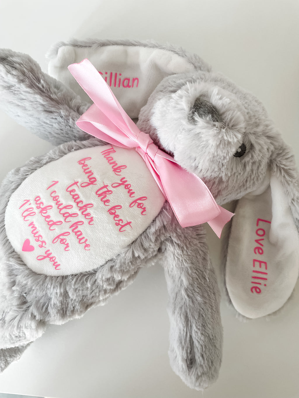 A personalised grey plush bunny with a pink satin bow, featuring personalised pink thank you teacher text on a white fabric belly patch and on the ear tabs, shown on a plain light background. Teacher Christmas gift.